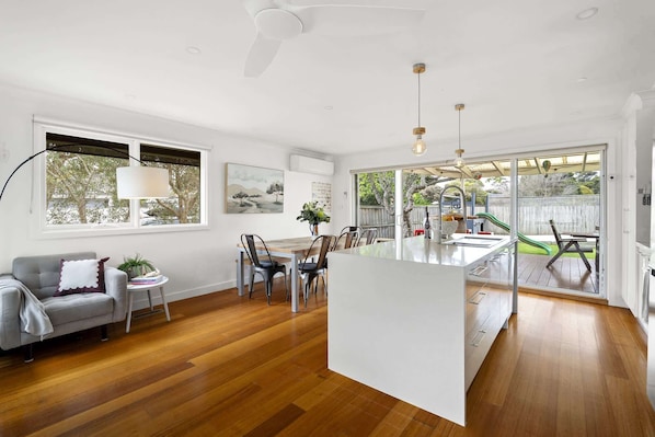 A modern kitchen with a central island, dining area with chairs, a sofa, and a large window.