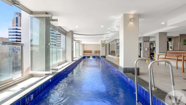 A long indoor swimming pool with clear blue water, surrounded by glass walls and a bench. - 2-Bed City Retreat with Gym and Pool Access (Adelaide)