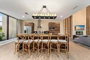 A modern kitchen with a central island, a dining area with wicker chairs, a large window, and a flat-screen TV mounted on a brick wall.
