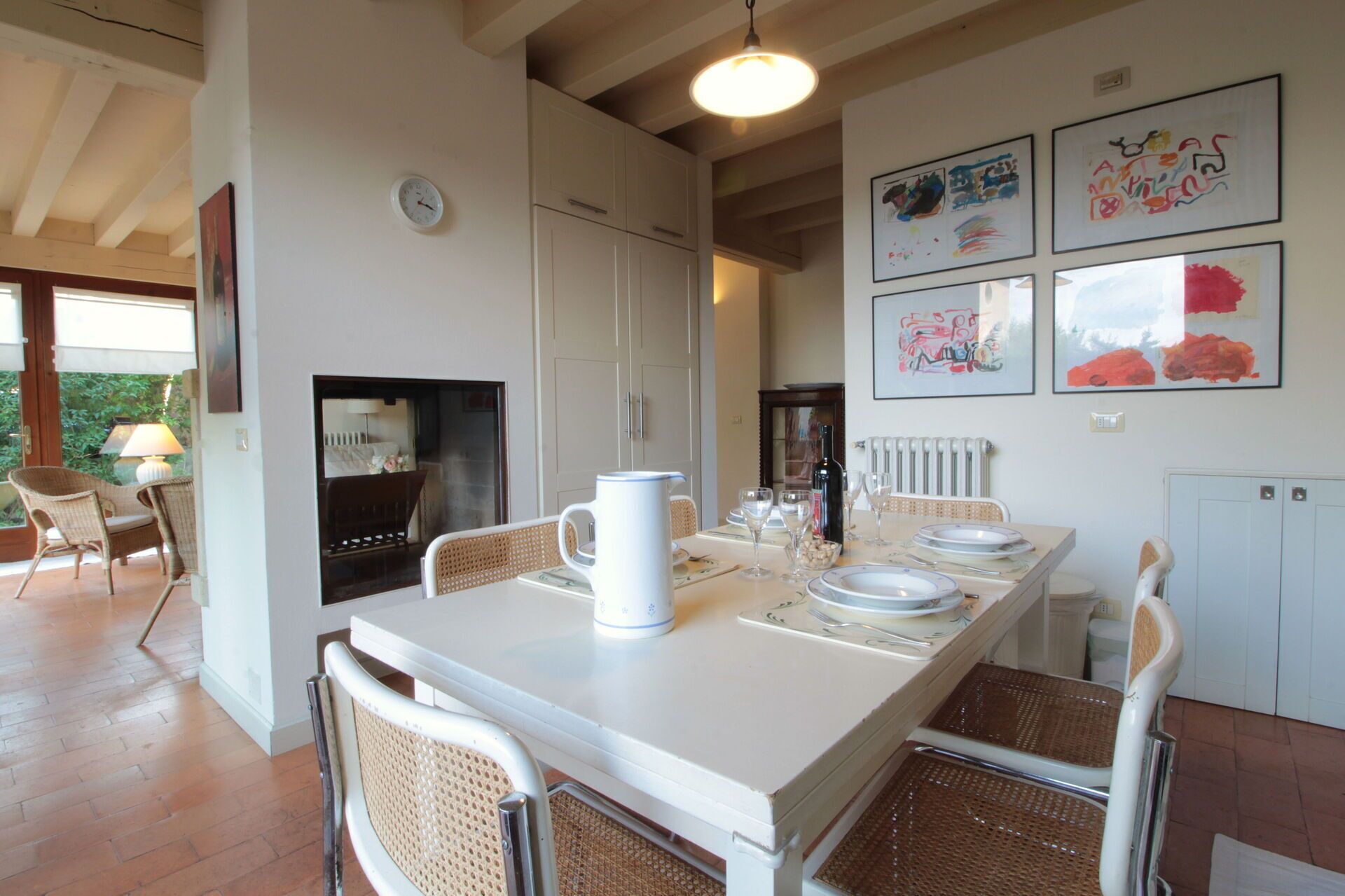 A dining area with a white table set for four, surrounded by wicker chairs. There are framed artworks on the wall above the table.