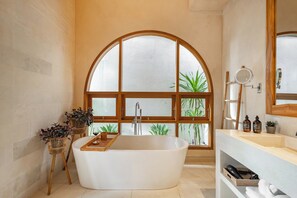 A modern bathroom with a freestanding bathtub, a large arched window, and a wooden vanity with a mirror and toiletries. - Villa Arkara Seminyak -- 3-bedroom villa, just 10-minutes walk to the beach (Bali)