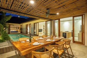 A dining area with a wooden table set for six, surrounded by wicker chairs, with a pool and a kitchen visible in the background. - Villa Arkara Seminyak -- 3-bedroom villa, just 10-minutes walk to the beach (Bali)