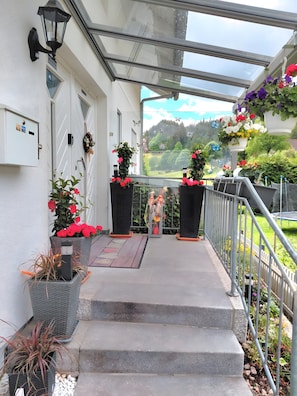 A covered outdoor area with steps leading to a door, potted plants, and a view of a green landscape.