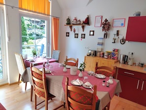 Un coin repas avec une table dressée pour quatre personnes, des chaises dotées de coussins à motifs, une fenêtre avec des rideaux et, au fond, des meubles de cuisine.