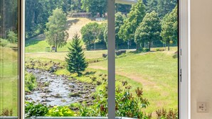 A view through a window of a natural landscape with a stream, trees, and a sign indicating 'Sandhill'. - Double Room 10 – with Shower, WC, Mountain & Stream View, Balcony, and Wi-Fi (Hornberg)