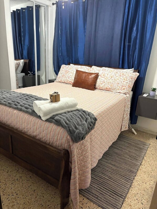 Un lit bien fait, avec une couvre-lit à motifs, des oreillers et une couverture. On y trouve une table de chevet avec une lampe et une petite plante.