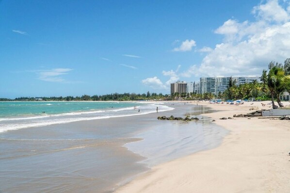Una playa con agua azul cristalina, orilla arenosa y una fila de edificios altos al fondo.