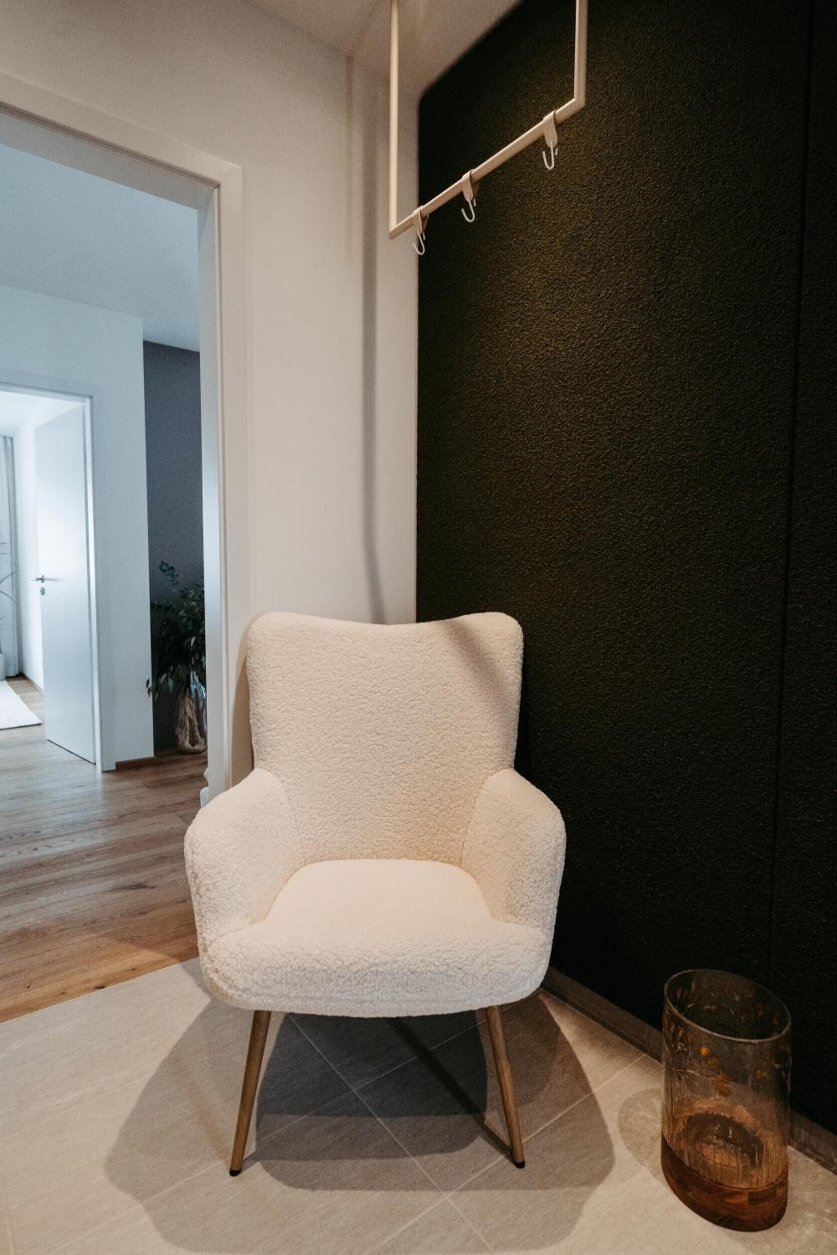 Un sillón blanco en una habitación con una pared oscura, una mesa de cristal con una bebida, y una puerta que da a otra habitación.