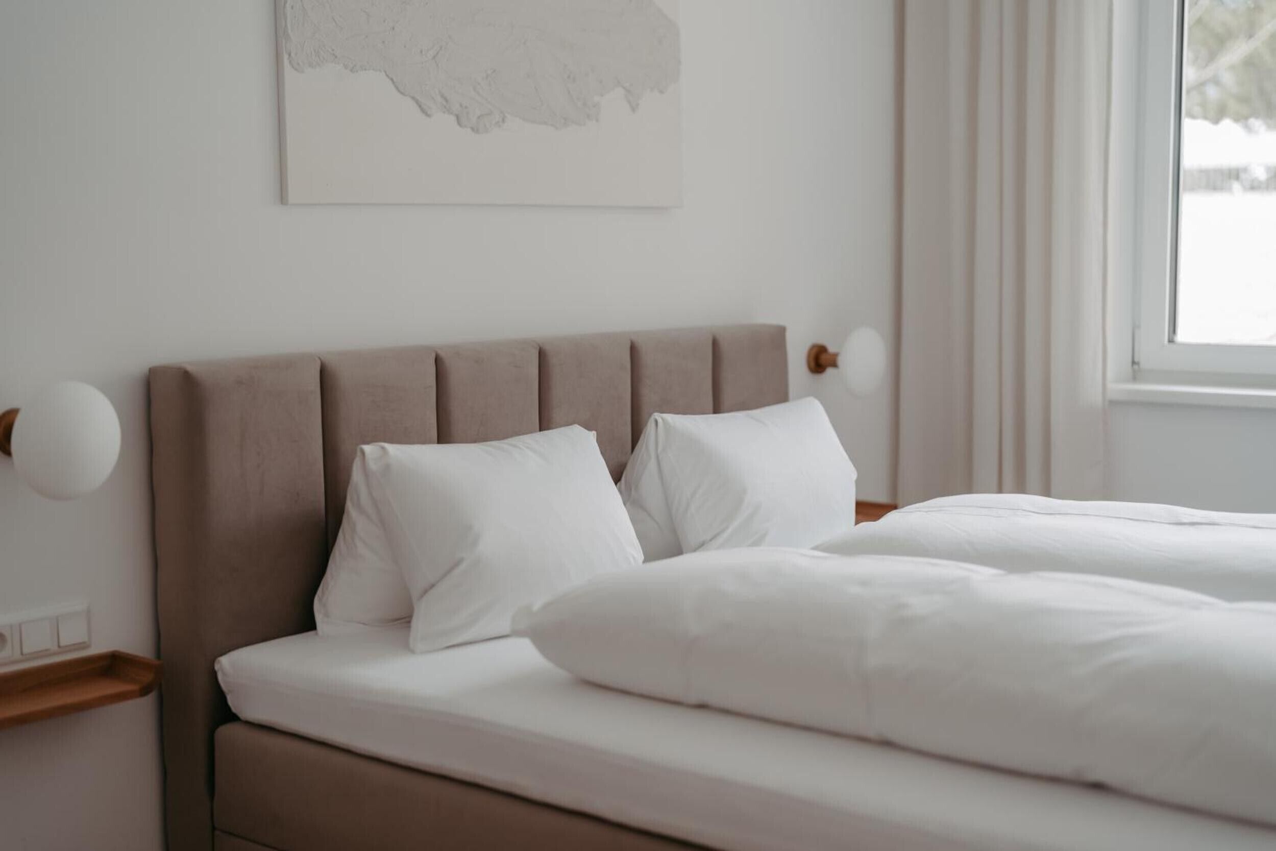 A neatly made bed with white linens and a headboard, a bedside table with a lamp, and a window with curtains.