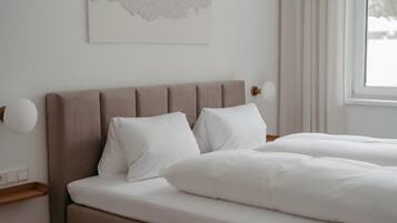 A neatly made bed with white linens and a headboard, a bedside table with a lamp, and a window with curtains.