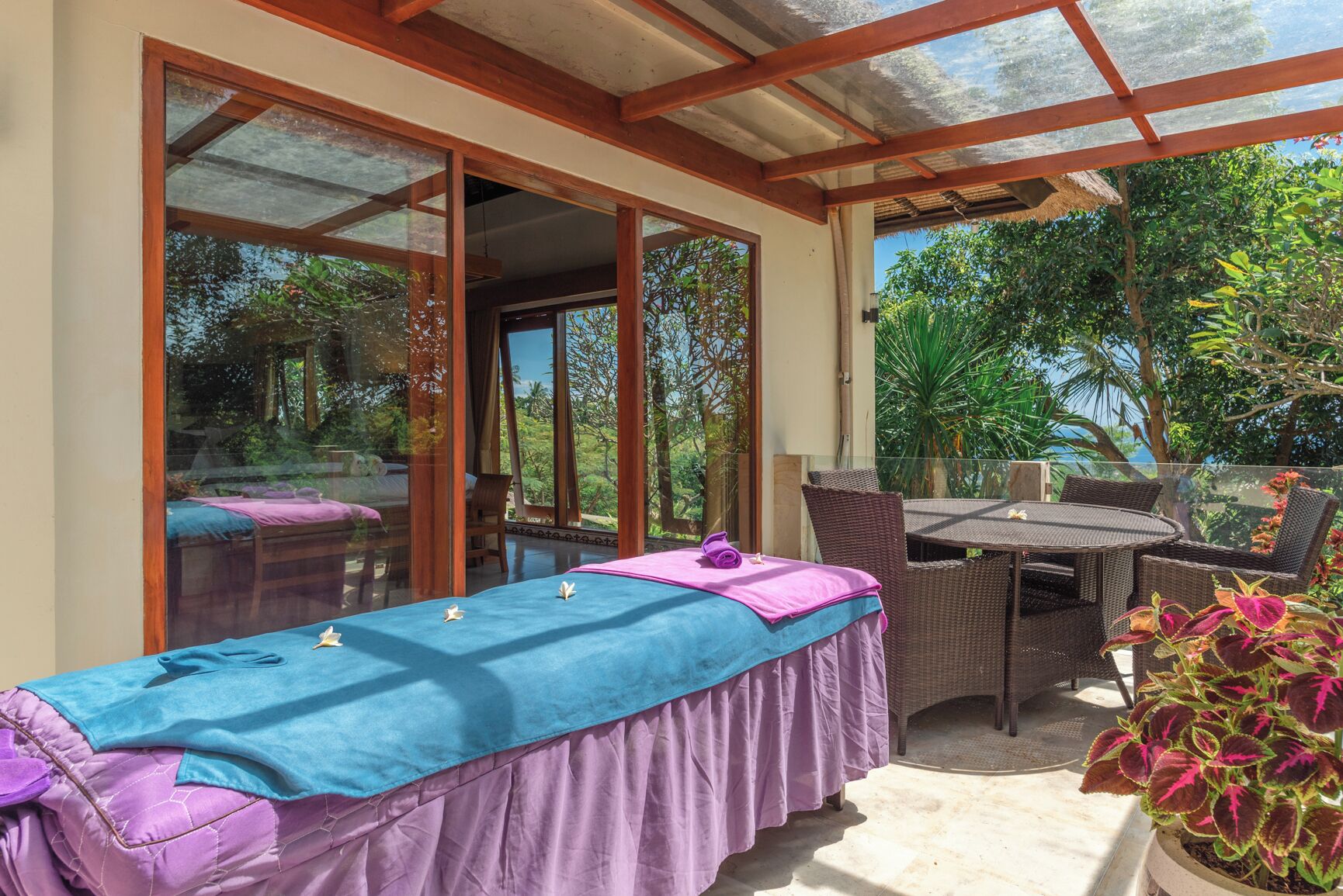 A room with a massage table, wicker chairs, and a glass-enclosed patio with a view of greenery.