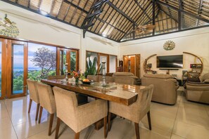 A spacious living area with a large dining table, comfortable seating, a television, and a view of the outdoors. - Bougainville Hill Villa (Sukasada)