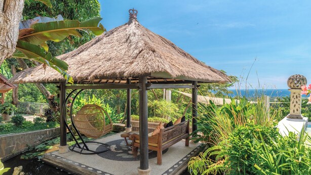 Ein Pavillon mit Strohdach und einer Bank, umgeben von üppiger Vegetation und Blick auf das Meer.