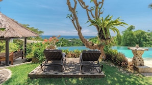 A poolside area with two lounge chairs facing a pool and ocean view, surrounded by lush greenery and a thatched-roof structure. - Bougainville Hill Villa (Sukasada)