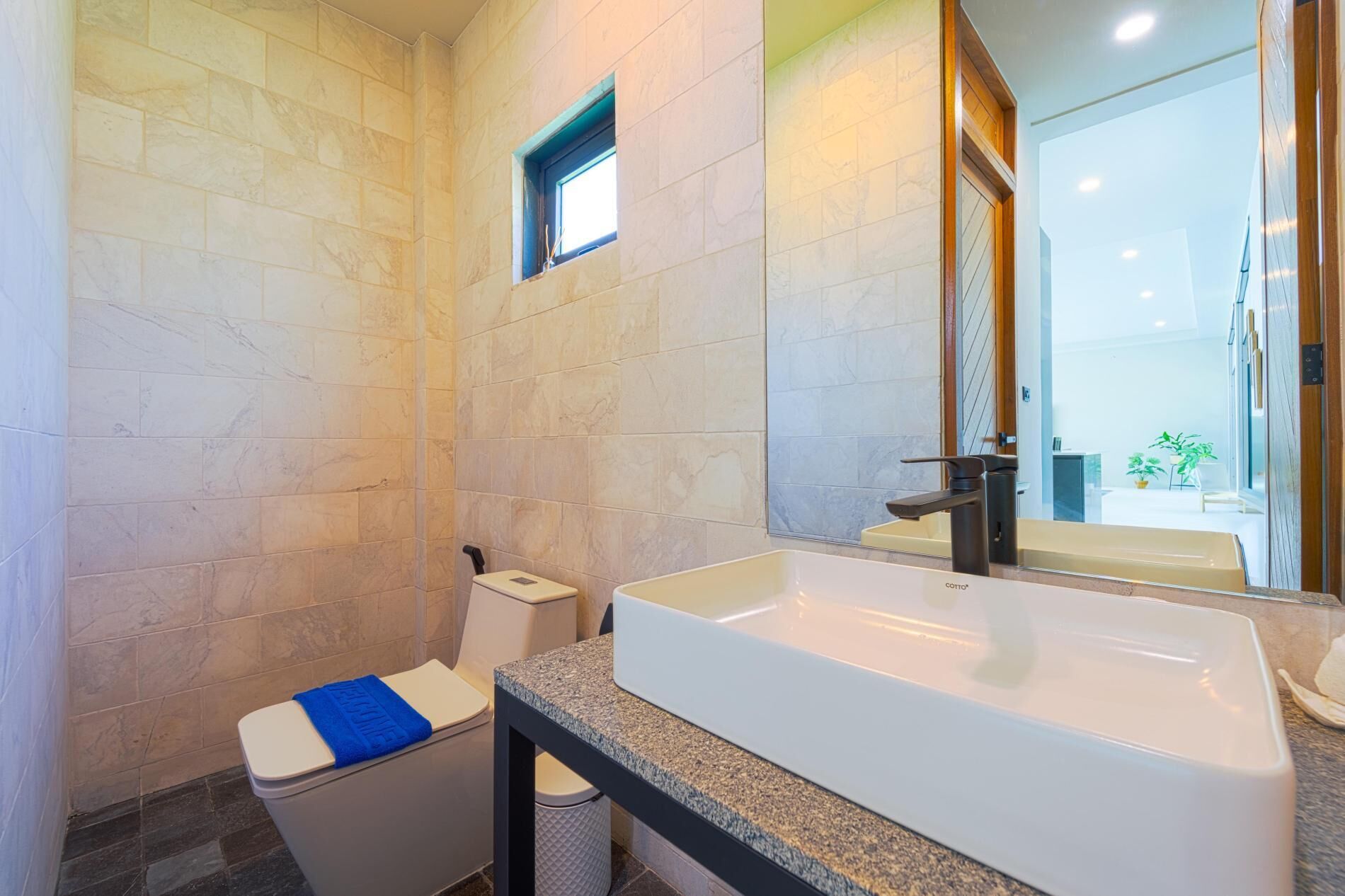 A modern bathroom with a large white sink, a toilet with a blue towel, and a small window.