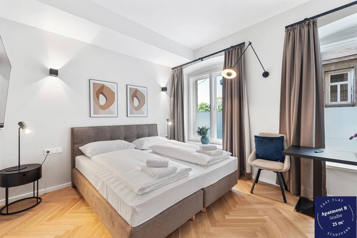 A modern bedroom with a double bed, bedside lamps, a desk with a chair, and a window with curtains.