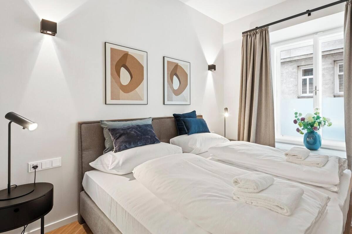 A modern bedroom with two beds, a bedside table with a lamp, and a window with curtains.