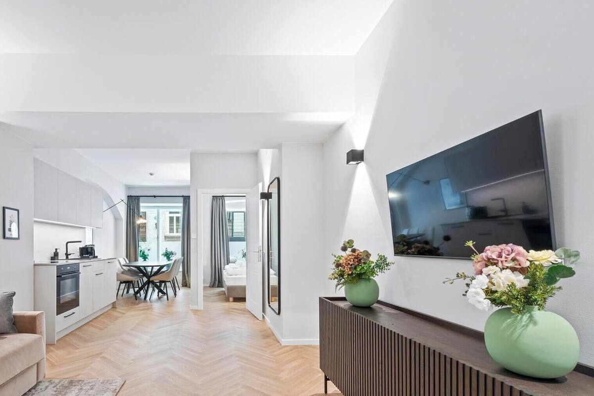A modern apartment with a kitchen, dining area, and living room. It features a herringbone wood floor, a sofa, a coffee table, and a TV mounted on the wall.