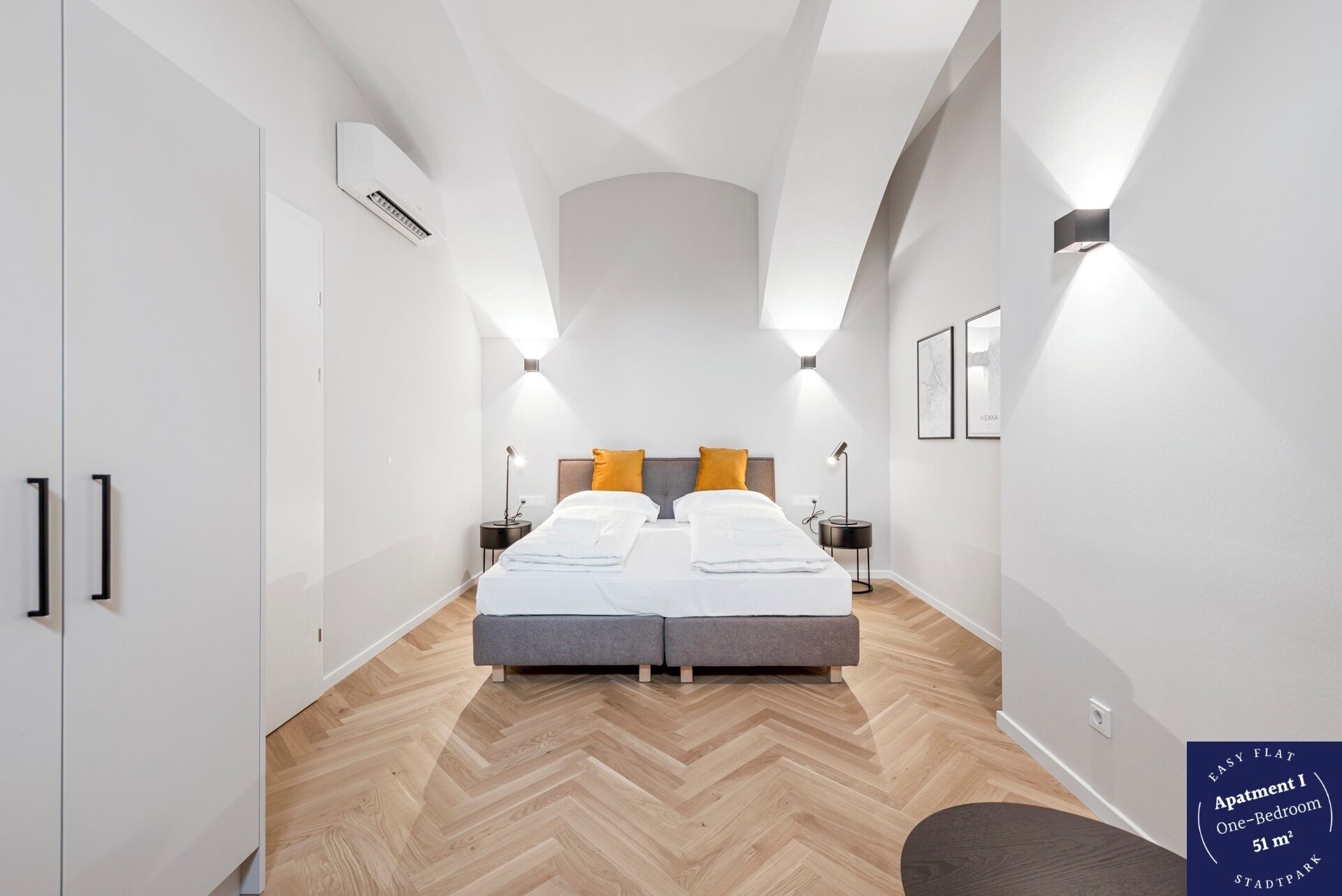 Eine moderne Einzimmerwohnung mit Fischgrätparkett, einem Bett mit weißer Bettwäsche und zwei orangefarbenen Kissen, einem Nachttisch mit Lampe sowie einer wandmontierten Klimaanlage.