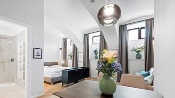 Una habitación de hotel moderna con cama, una mesa de comedor y zona de estar. Hay un ventanal con cortinas, un área de ducha y un florero con flores sobre la mesa.