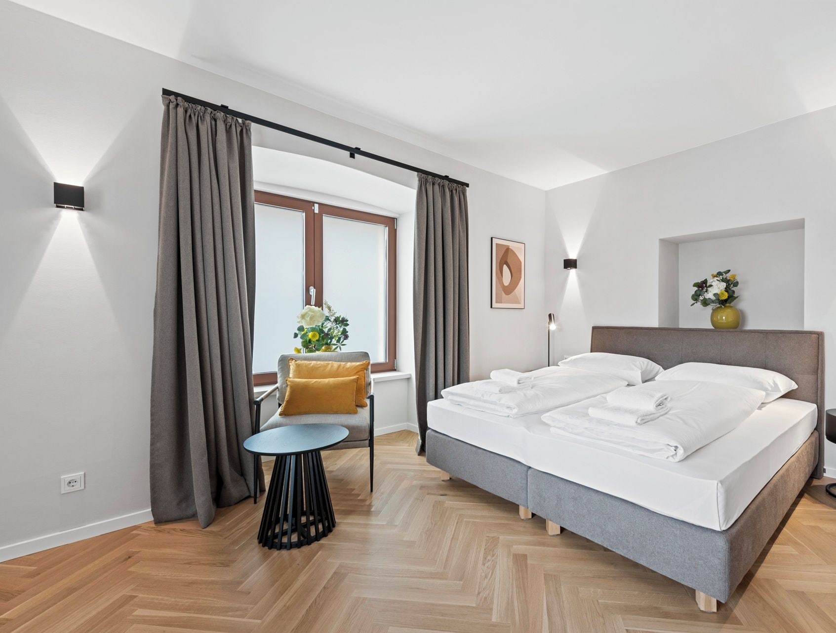 A modern bedroom with a large bed, a small round table, and a chair. There is a window with curtains, a wall-mounted lamp, and a framed picture.