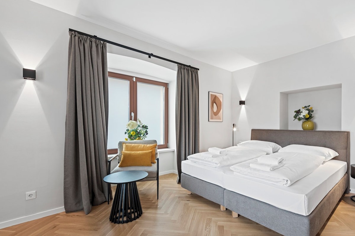 A modern bedroom with a large bed, a small round table, and a chair. There is a window with curtains, a wall-mounted lamp, and a framed picture.