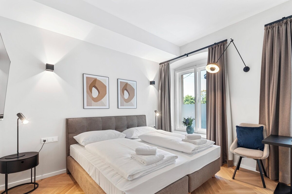 A modern bedroom with a double bed, bedside lamps, a desk with a chair, and a window with curtains.