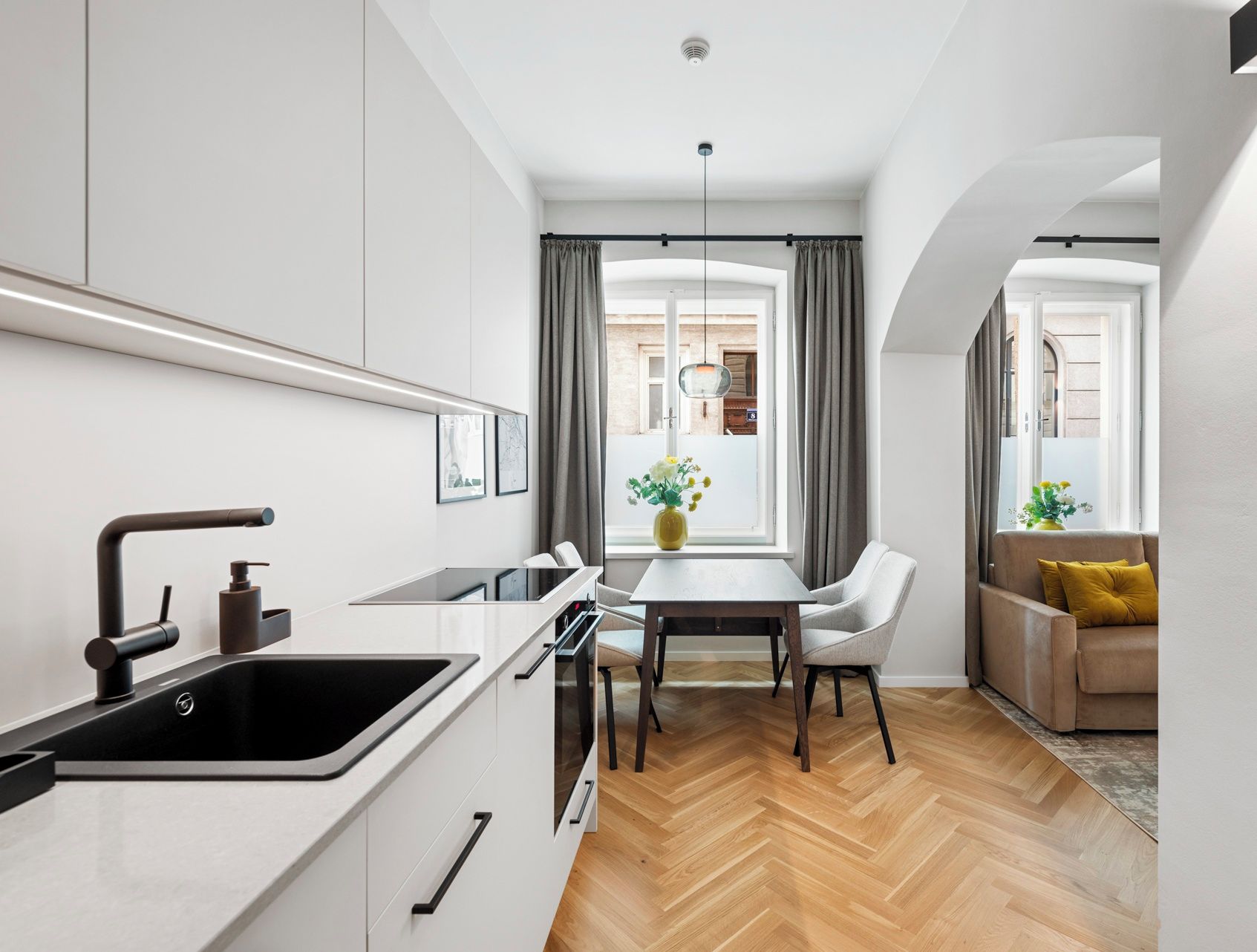 Una cocina moderna con un lavamanos, estufa y zona de comedor. Hay una ventana con cortinas, un sofá y una mesa con sillas.
