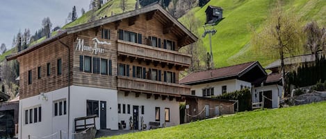 A two-story lodge with a wooden facade and a balcony, set against a backdrop of green hills and a clear sky.
