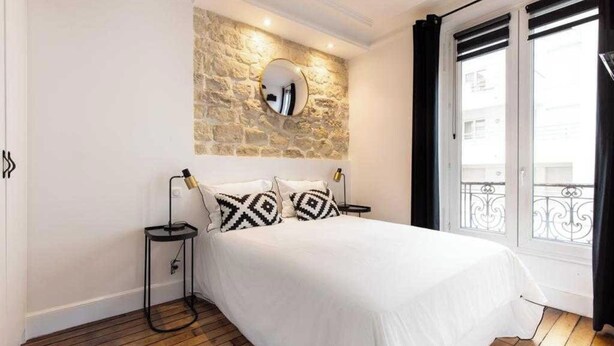 A bedroom with a stone accent wall, a bed with patterned pillows, a bedside lamp, and a view of a balcony. - Spacious Apartment 2BR/5P – Nogent-sur-Marne (Nogent-sur-Marne)