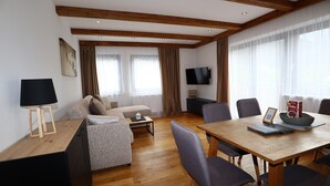 En moderne stue med sofa, sofabord, spisebord med stole, et fjernsyn og et stort vindue med gardiner.
