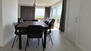 Un espace repas moderne avec une table rectangulaire et six chaises, de grandes fenêtres avec des rideaux et une vue sur l’extérieur.