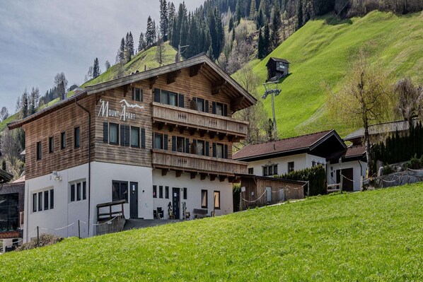 A two-story lodge with a wooden facade and a balcony, set against a backdrop of green hills and a clear sky.