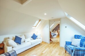 Une chambre spacieuse aménagée en loft, avec un plafond haut, des fenêtres de toit, un plancher en bois, un canapé blanc, un canapé à carreaux bleus et un lit dont la tête de lit est recouverte d’un papier peint à motifs floraux.