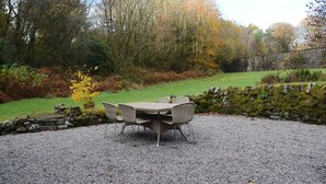 An outdoor seating area with a table and chairs, surrounded by a gravel ground, with trees and a stone wall in the background.