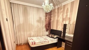 A bedroom with a wooden floor, a bed with a white and red patterned bedspread, a black headboard, a nightstand, a dresser, and a chandelier. - Sabir bir (Baku)
