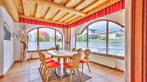 Een eetruimte met een houten tafel en stoelen, grote ramen met gordijnen en uitzicht op huizen en bergen.