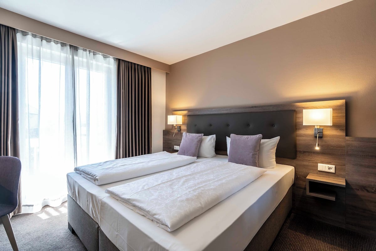 Ein Hotelzimmer mit zwei Betten, einem Schreibtisch, einem Sessel, einem Nachttisch mit Lampe und einem Fenster mit Vorhängen.