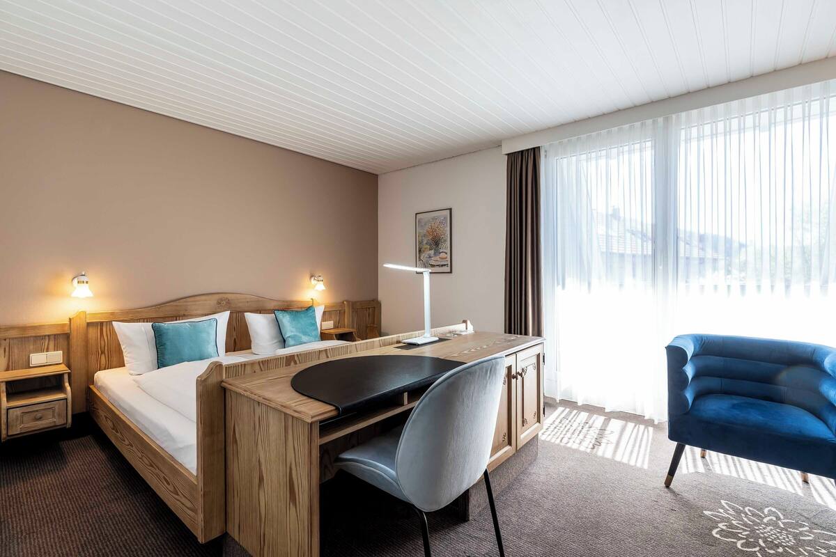 Ein Hotelzimmer mit Bett, Schreibtisch, Stuhl und einem großen Fenster mit Vorhängen.