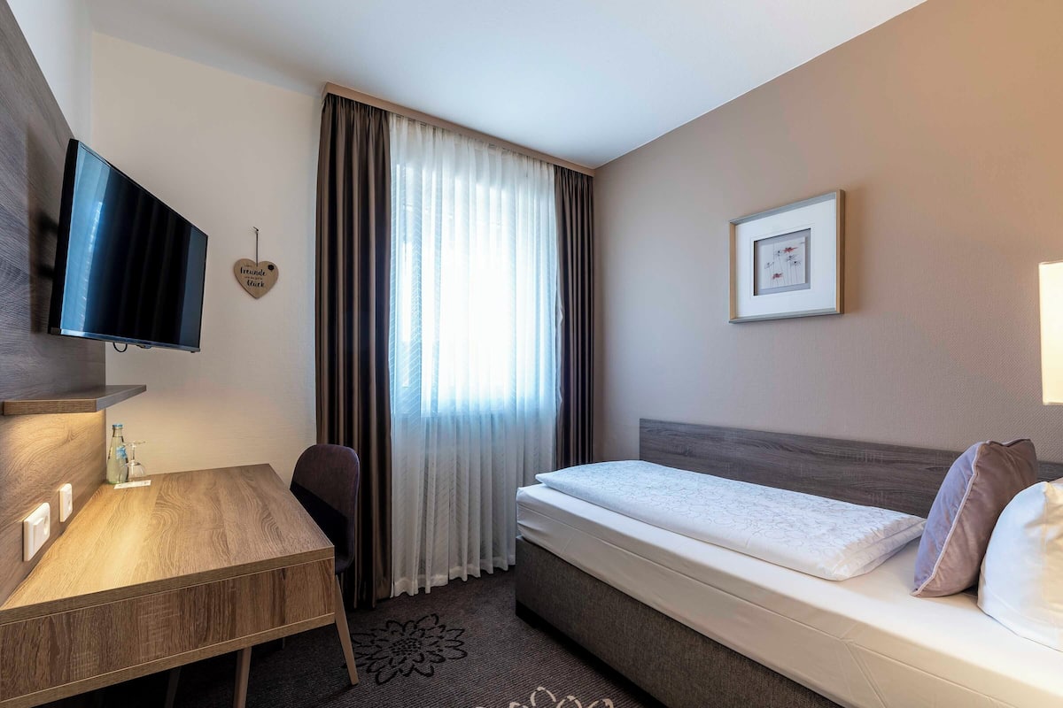 Ein Hotelzimmer mit Bett, Schreibtisch, Stuhl, Fernseher und einem Fenster mit Vorhängen.