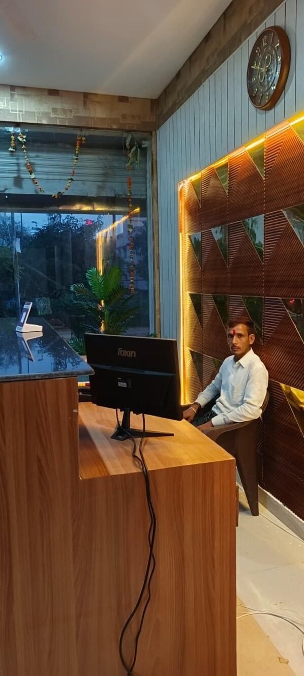 A reception desk with a computer, a clock on the wall, and a window with a view of the outside. - Swastik Inn (Gwalior)