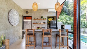 A kitchen with a bar area, wooden chairs, hanging wicker lights, a large decorative plate, and a view of a swimming pool. - Villa Indra Pererenan by Orivista (Canggu)