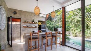 A modern kitchen with a bar area, large windows, and a view of a swimming pool. - Villa Indra Pererenan by Orivista (Canggu)