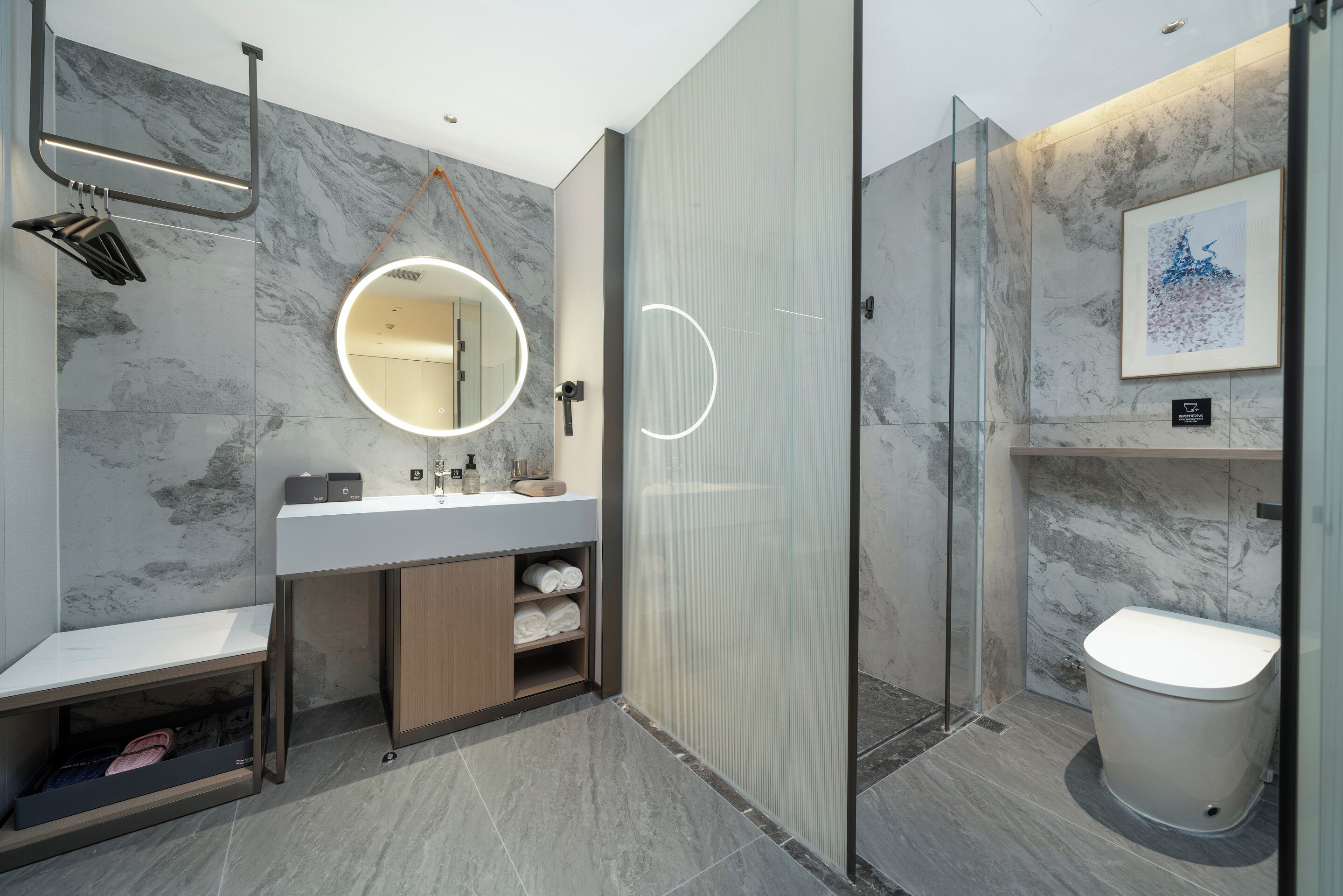 Un baño moderno con salpicadero de mármol, un espejo redondo con luz, cabina de ducha de vidrio y un inodoro.