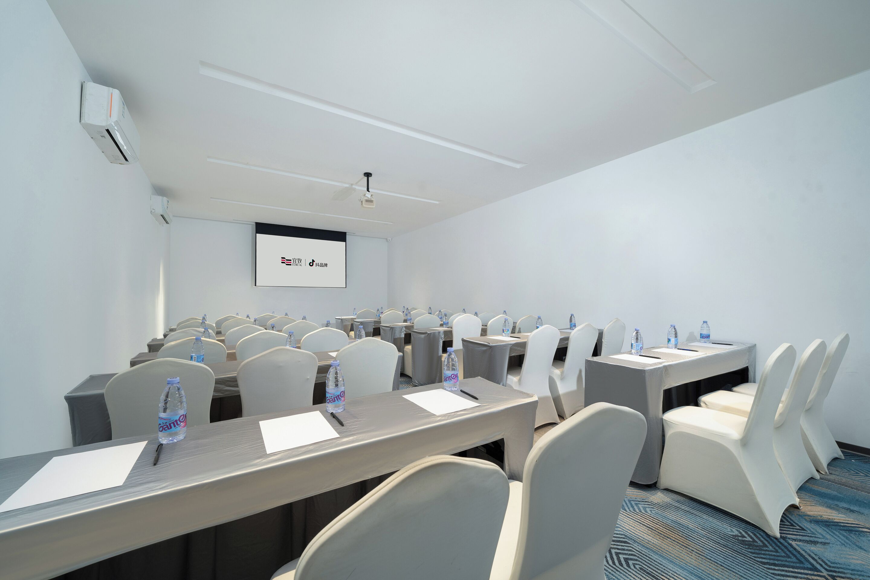Un salón de conferencias con filas de mesas y sillas, una pantalla de proyector y botellas de agua sobre las mesas.
