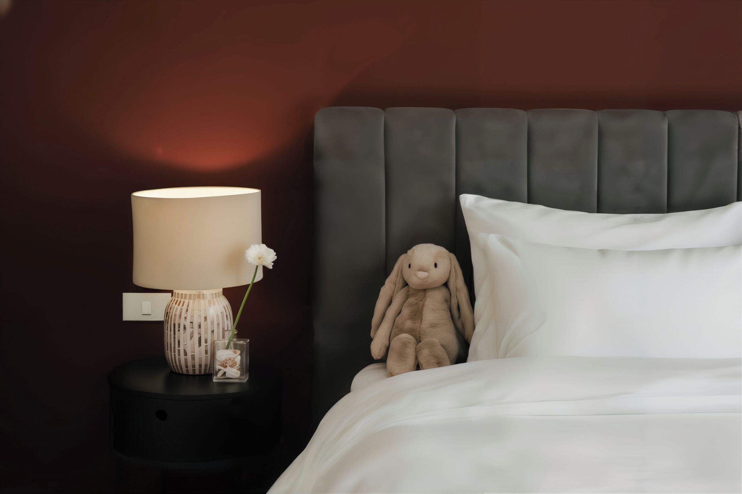 A neatly made bed with a plush headboard, a bedside lamp, and a small vase with a flower.