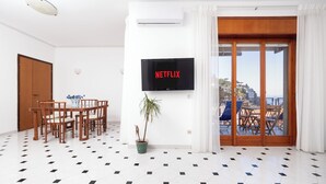 A room with a dining table and chairs, a potted plant, a TV mounted on the wall, and a sliding glass door leading to an outdoor area with a view of a hill and the sea. - Capo Sereno (Sorrento)