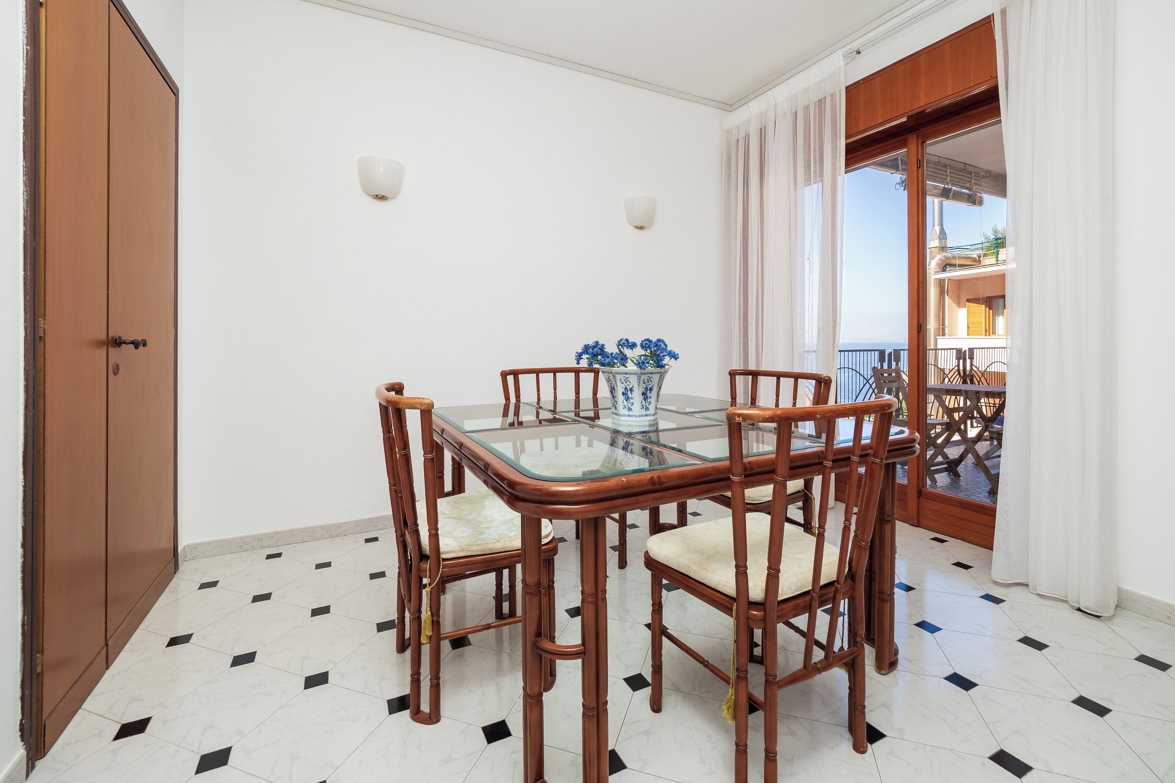A dining area with a glass-top table, four wooden chairs, a vase with flowers, and a view of a balcony with a railing.