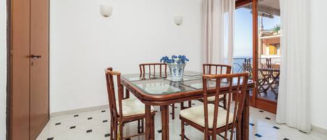 A dining area with a glass-top table, four wooden chairs, a vase with flowers, and a view of a balcony with a railing.
