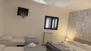 A hotel room with three beds, a flat-screen TV, a window with blinds, a radiator, and a patterned wallpaper. - An elegant retreat overlooking the city of mosaics (Caltagirone)
